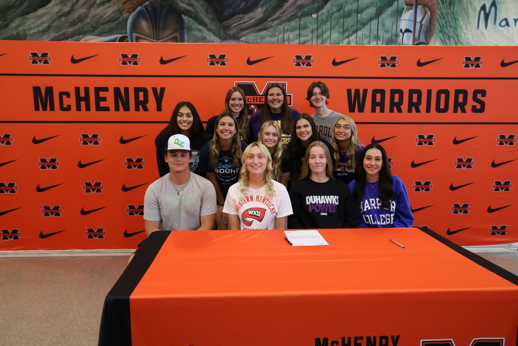 Twelve students at the signing table with. McHenry Warriors backdrop